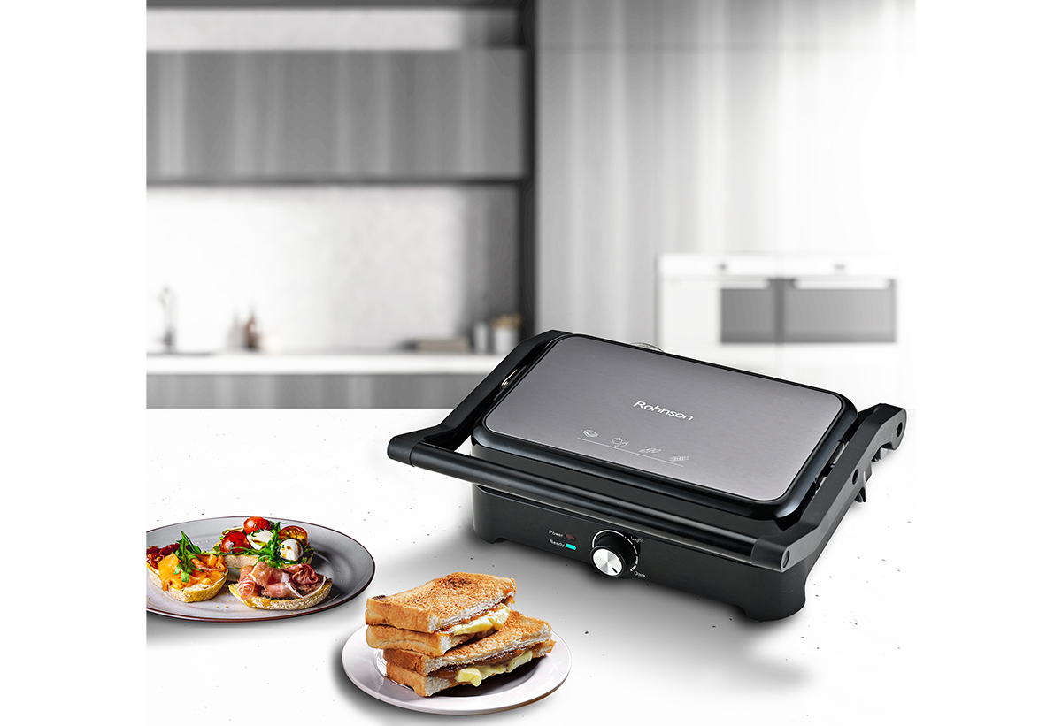 Απεικονίζεται η τοστιέρα δίπλα σε 2 πιάτα από τα φαγητά που έχει παρασκευάσει.